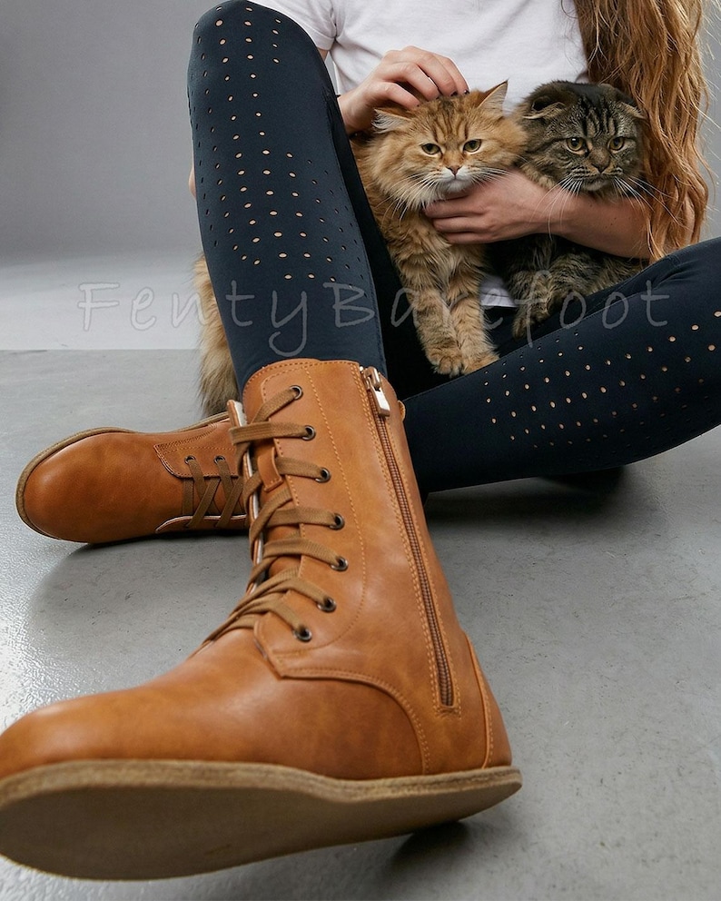 Op de afbeelding: Lichtbruine veterlaarzen met een zijrits. De laarzen worden gedragen met donkerblauwe leggings met kleine cirkelvormige uitsparingen. Twee katten worden op schoot gehouden. De tekst "FentyBarefoot" is zichtbaar.