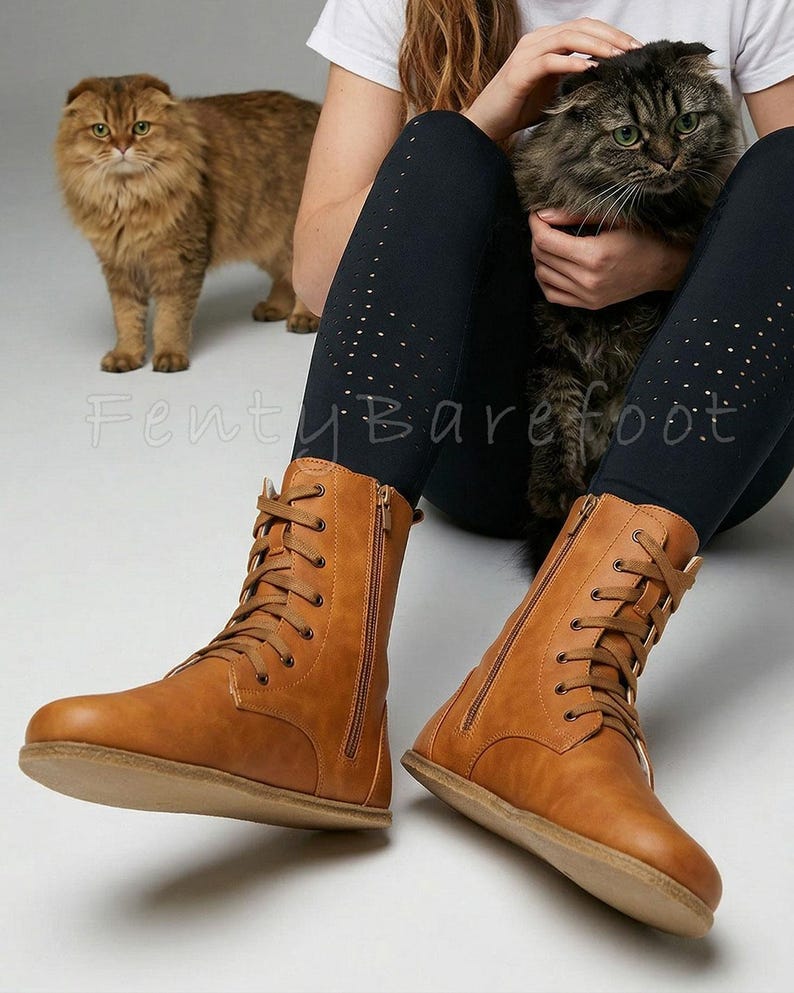 Op de afbeelding: Een paar bruinleren veterlaarzen met zijritsen. De laarzen hebben een platte zool en zijn gemaakt van glad materiaal. De laarzen worden getoond met een zwarte legging en twee katten op de achtergrond.
