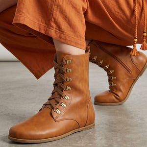 May include: A pair of brown leather lace-up boots with gold-colored eyelets and a side zipper. The boots have a flat sole and are worn with orange pants. The Etsy shop name "fentybarefoot.etsy.com" is visible at the bottom of the image.