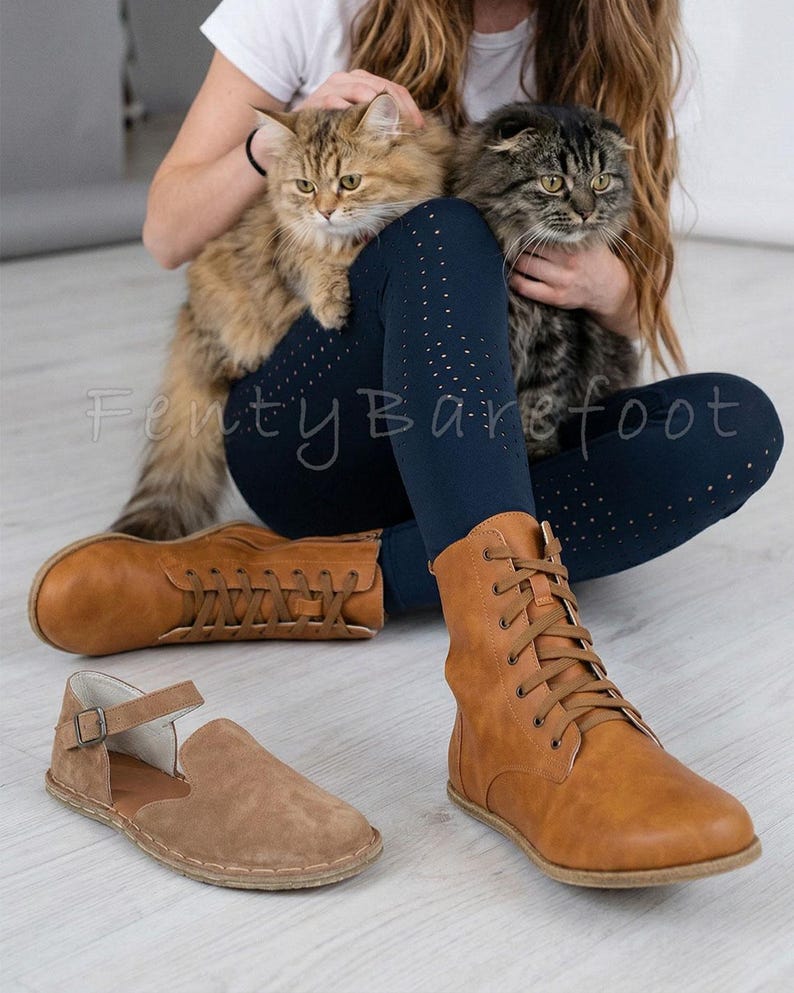 Op de afbeelding: Er worden twee paar schoenen getoond: bruine leren laarzen en bruine su&egrave;de sandalen. De laarzen zijn geregen en hoog, terwijl de sandalen een bandje en een gesp hebben. De tekst "Fenty Barefoot" is zichtbaar.