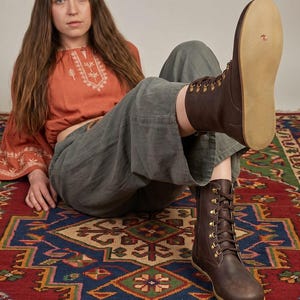 May include: Brown leather lace-up boots with brass eyelets and a light-colored sole. The boots are worn with gray pants and a rust-colored top with white embroidery. The boots are a key element in this fashion-focused image.