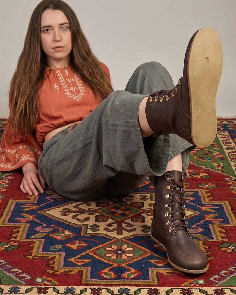 May include: Brown leather lace-up boots with brass eyelets and a light-colored sole. The boots are worn with gray pants and a rust-colored top with white embroidery. The boots are a key element in this fashion-focused image.
