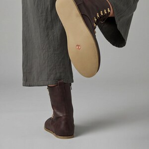 May include: A pair of brown leather boots with a flat sole. The boots have a mid-calf height and feature gold-colored eyelets and laces. The sole is a light tan color. The boots are shown with a person wearing gray pants.