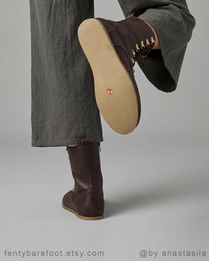 May include: A pair of brown leather boots with a flat sole. The boots have a mid-calf height and feature gold-colored eyelets and laces. The sole is a light tan color. The boots are shown with a person wearing gray pants.