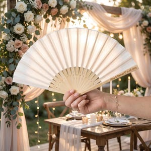 May include: A white paper fan with a light wood handle is held in front of a wedding arch. The fan has the text "Nina & Itamar 21.08.2025" printed on the handle. The background includes flowers, candles, and a table setting.
