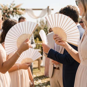 Puede incluir: Varias personas sostienen abanicos de papel blanco abiertos con mangos de madera. Los abanicos se sostienen en alto. Los abanicos tienen un diseño plisado. El fondo incluye un arco de boda y otros invitados.