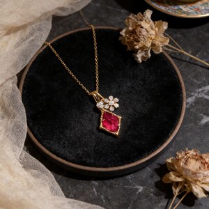 Könnte beinhalten: Eine goldene Halskette mit einem Anhänger, der einen großen, roten Edelstein in Diamantenform aufweist, akzentuiert durch eine Ansammlung kleiner, klarer Steine. Die Halskette liegt auf einer schwarzen Samtoberfläche, mit getrockneten Blumen und einer gemusterten Teetasse im Hintergrund.