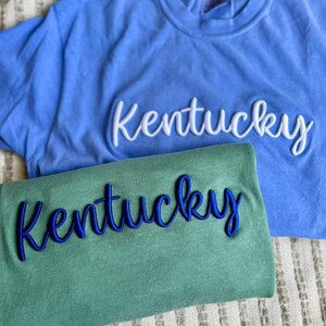 May include: Two t-shirts are shown, one blue and one green. Each shirt features the word "Kentucky" embroidered in a cursive style. The blue shirt has white embroidery, and the green shirt has blue embroidery.