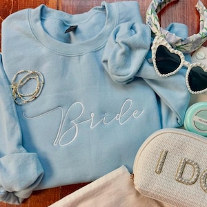 Puede incluir: Una sudadera azul claro con la palabra "Bride" bordada en escritura blanca. La imagen también incluye una pulsera de perlas, gafas de sol en forma de corazón, una diadema floral y un neceser blanco con "I DO" en letras de perlas.