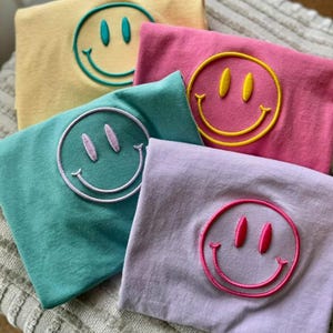 May include: Four folded t-shirts in pastel colors including yellow, pink, teal, and lavender. Each shirt features a large, centered, embroidered smiley face icon in a contrasting thread color, specifically teal, yellow, white, and hot pink.