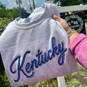 Może przedstawiać: Jasnoszara bluza z okrągłym dekoltem, na której wyhaftowano słowo Kentucky pogrubioną, kursywną niebieską czcionką z ciemnoniebieskim obrysem. Odzież posiada klasyczny prążkowany dekolt oraz długie rękawy, prezentując prosty i czysty design.