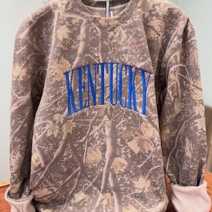 May include: A camouflage-patterned sweatshirt with the word "KENTUCKY" embroidered in blue across the chest. The sweatshirt has a crew neck and long sleeves with white cuffs. The fabric design resembles a forest with brown and tan tones.