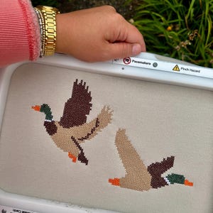 May include: A white rectangular embroidery hoop containing light beige fabric with a cross-stitch pattern of two flying mallard ducks. The ducks feature brown, tan, green, and orange thread. The hoop frame includes safety warning text labels.