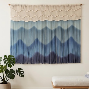 May include: A large, textured wall hanging with a gradient of blues and a cream-colored top section. The design features a wave-like pattern and is mounted on a wooden rod. The piece is displayed in a room with a plant and a bench.