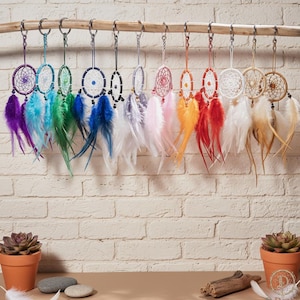May include: A display of dream catchers in a range of colours suspended from a wooden branch. Each dream catcher has a circular woven web design, with feathers in shades of purple, blue, green, and orange. They are hung by silver chains.