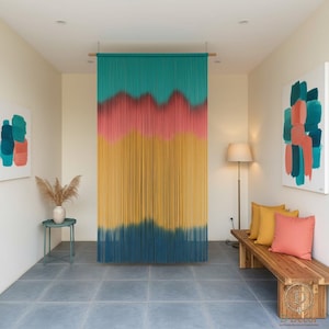 Könnte beinhalten: Ein farbenfroher, handgefertigter Vorhang hängt in einem Türrahmen. Der Vorhang hat ein Farbverlauf-Design mit Türkis-, Rosa-, Gelb- und Marineblautönen. Er besteht aus vielen Materialsträngen, wodurch ein strukturierter, geschichteter Effekt entsteht. Der Vorhang ist an einer Holzstange aufgehängt.