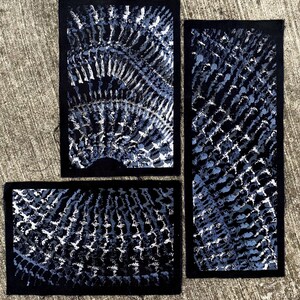 Könnte beinhalten: Drei rechteckige Stoffaufnäher mit einer tiefschwarzen Basis und einem sich wiederholenden, strukturierten Muster in Blau-, Grau- und Weißtönen. Das Design besteht aus geschwungenen, strahlenförmigen Linien, die Tiefe erzeugen.
