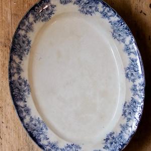 Könnte beinhalten: Eine ovale Servierplatte aus Keramik mit cremefarbener Mitte und einem breiten Rand, der mit einem detaillierten dunkelblauen Blumenmuster verziert ist. Die Ränder der Platte sind gewellt und die Oberfläche weist Altersspuren auf.