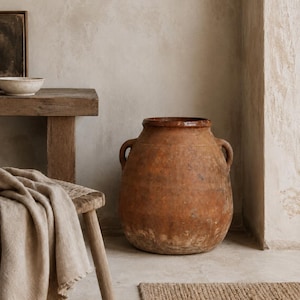 May include: A large, weathered terracotta vessel with a wide mouth and two side handles. The pot features a rustic, uneven surface with mottled brown and orange tones, showing signs of age and texture throughout its rounded, bulbous body.
