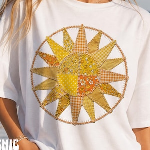 May include: A white short-sleeved t-shirt featuring a large circular sun graphic on the chest. The sun is composed of various yellow and orange fabric-style patches with floral, polka dot, and gingham patterns, outlined with a stitched border.