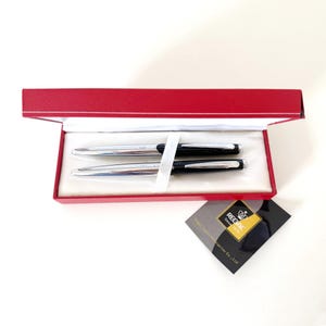 Op de afbeelding: Een set van twee pennen in een rode presentatiedoos. Eén pen is zilverkleurig, de andere zwart. De pennen liggen op een wit, satijnachtig materiaal. Een klein label met de tekst "REGAL" is zichtbaar.
