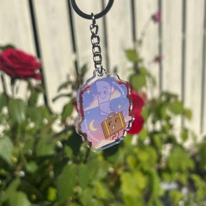 May include: A clear acrylic keychain featuring a cartoon illustration of a knife and suitcase with smiling faces. The background shows a night sky with a crescent moon and stars. The keychain is attached to a silver chain and ring.