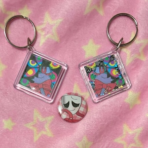 May include: Two square acrylic keychains featuring a colorful character with a red bow tie and a circular pinback button displaying a white mask with closed eyes and a red ribbon, all arranged together on a soft pink fabric surface.