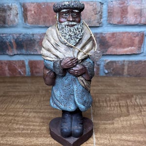 Può includere: Statuetta dettagliata di un uomo con una lunga barba bianca, che indossa un cappotto blu, un cappello e uno scialle beige. La statuetta è su una base a forma di cuore. Una piccola etichetta con la scritta "GREEN LANE" è appesa.