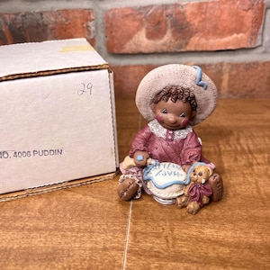 May include: A ceramic figurine of a child wearing a hat and a pink dress, holding a cake that says "Happy Birthday." A small teddy bear sits next to the child. The figurine is on a wooden surface, with a cardboard box in the background.