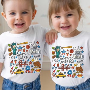 May include: A white short-sleeved t-shirt featuring a colorful graphic print of American icons including the Statue of Liberty, a bald eagle, the Capitol building, a taxi, and fireworks, with the text 1776 HAPPY 250 AMERICA 2026.