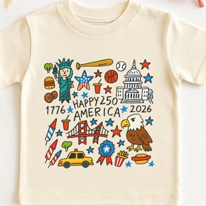 May include: A natural-colored short-sleeve t-shirt featuring a colorful graphic print of American icons including the Statue of Liberty, a bald eagle, the Capitol building, a taxi, and the text Happy 250 America 1776-2026, with pink flower-shaped sunglasses.