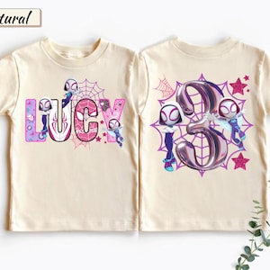 K&ouml;nnte beinhalten: Cremefarbenes T-Shirt mit dem Namen "Lucy" in einer farbenfrohen Comic-Schriftart, mit Spider-Gwen-Charakteren und einem Spinnennetz-Design. Die R&uuml;ckseite zeigt eine gro&szlig;e "3" mit Spider-Gwen-Charakteren und Sternen.