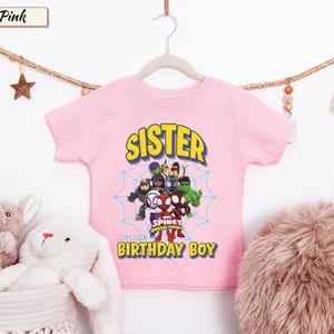 May include: A light pink short-sleeved t-shirt featuring a graphic of Marvel Spidey and His Amazing Friends characters centered within a blue spiderweb. The text reads SISTER OF THE BIRTHDAY BOY in yellow block letters with a dark blue outline.