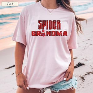 May include: A light pink t-shirt with the words "SPIDER GRANDMA" in red, distressed lettering. A small spider graphic is between the words. The shirt has short sleeves and a relaxed fit.