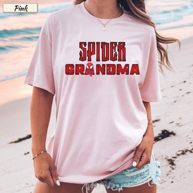 May include: A light pink t-shirt with the words "SPIDER GRANDMA" in red, distressed lettering. A small spider graphic is between the words. The shirt has short sleeves and a relaxed fit.