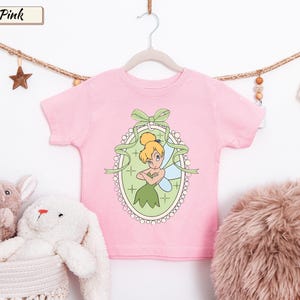 May include: A light pink short-sleeved t-shirt featuring a graphic of a blonde fairy with wings inside an oval frame decorated with a green bow. The design includes a light green background with small star accents and a white beaded border.
