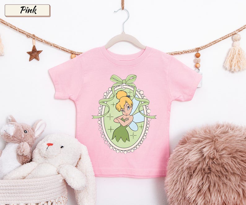 May include: A light pink short-sleeved t-shirt featuring a graphic of a blonde fairy with wings inside an oval frame decorated with a green bow. The design includes a light green background with small star accents and a white beaded border.