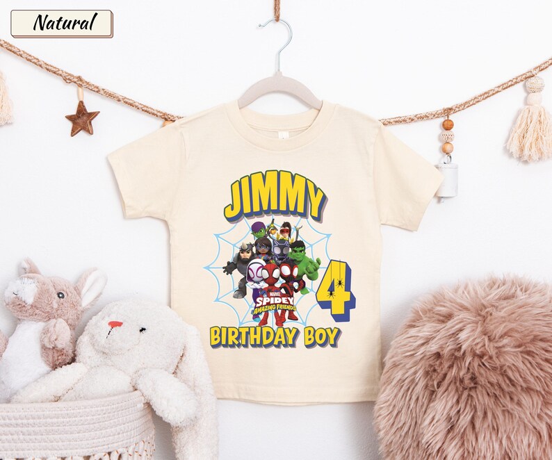 May include: A natural-colored short-sleeve t-shirt featuring a graphic of Marvel Spidey and His Amazing Friends characters, a blue spiderweb, the name JIMMY, the number 4, and the text BIRTHDAY BOY in yellow and blue lettering.