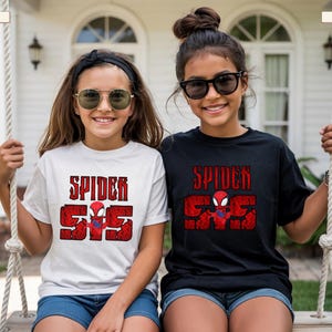 May include: Two girls wearing Spider-Man t-shirts. One shirt is white, the other is black. Both shirts have the words "SPIDER" and "555" in red, with a Spider-Man graphic. The girls are smiling and wearing sunglasses.