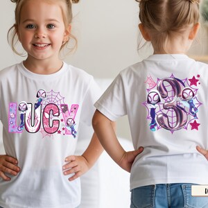 K&ouml;nnte beinhalten: Wei&szlig;es T-Shirt mit dem Namen "LUCY" in rosa und lila Buchstaben, Spinnennetz-Design und Cartoon-Figuren. Auf der R&uuml;ckseite des Shirts ist eine gro&szlig;e "3" mit Spinnennetz- und Sternakzenten abgebildet.