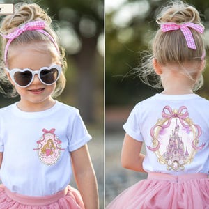 Op de afbeelding: Een wit t-shirt met korte mouwen en een kleine afbeelding op de linkerborst van een prinses in een gouden ovaal frame met een roze strik. De achterkant van het shirt toont een grotere versie van hetzelfde gouden frame met een kasteel.
