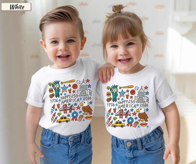 May include: A white short-sleeved t-shirt featuring a colorful graphic print of American icons including the Statue of Liberty, a bald eagle, the Capitol building, a taxi, and fireworks, with the text 1776 HAPPY 250 AMERICA 2026.