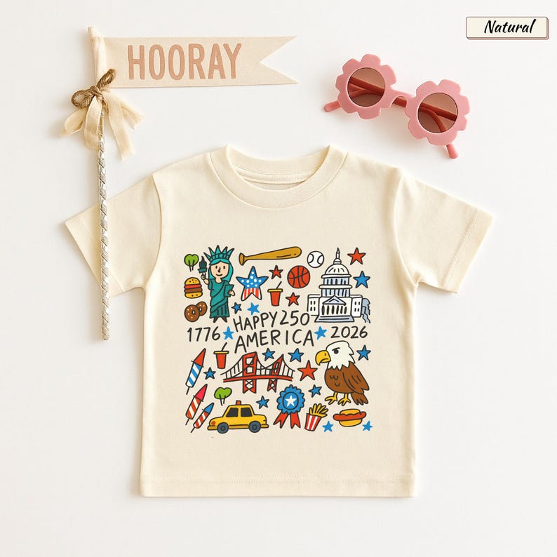 May include: A natural-colored short-sleeve t-shirt featuring a colorful graphic print of American icons including the Statue of Liberty, a bald eagle, the Capitol building, a taxi, and the text Happy 250 America 1776-2026, with pink flower-shaped sunglasses.