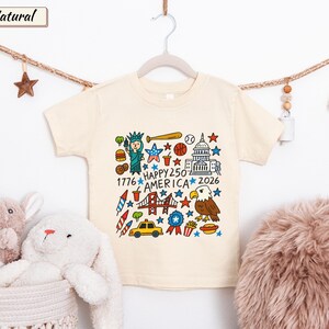 May include: A natural-colored short-sleeve t-shirt featuring a colorful graphic print of American icons including the Statue of Liberty, the Capitol building, a bald eagle, and a taxi, with the text "HAPPY 250 AMERICA 1776 2026" in the center.