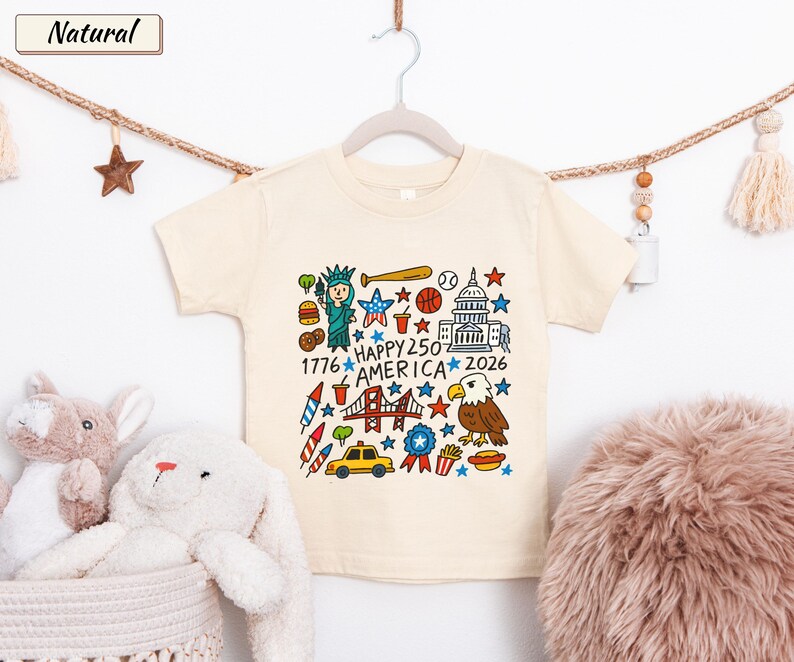 May include: A natural-colored short-sleeve t-shirt featuring a colorful graphic print of American icons including the Statue of Liberty, the Capitol building, a bald eagle, and a taxi, with the text "HAPPY 250 AMERICA 1776 2026" in the center.