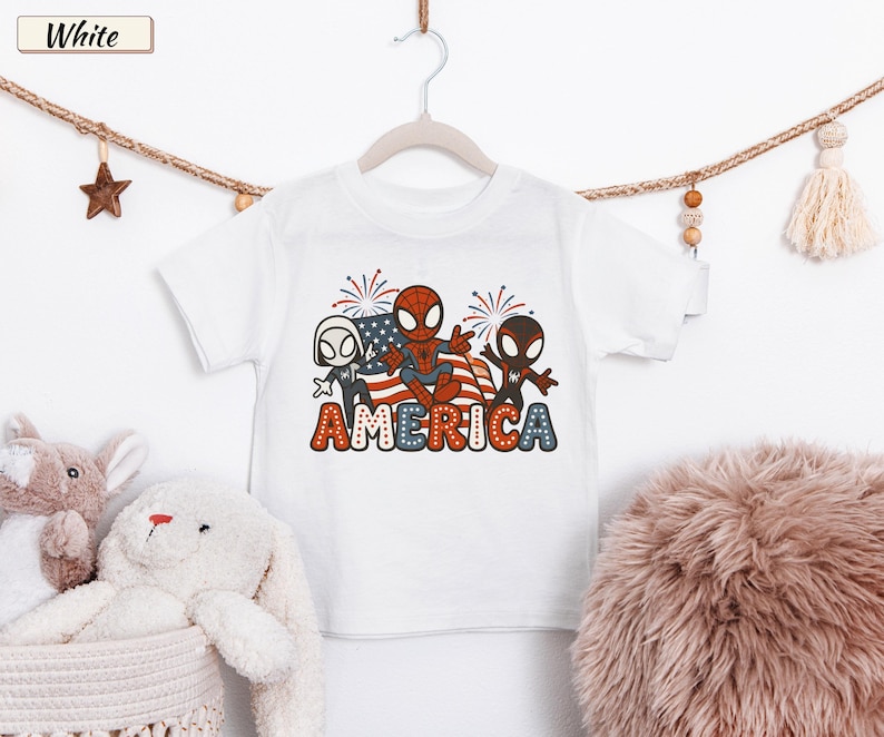 May include: A white short-sleeved t-shirt featuring a graphic of three cartoon spider-themed characters in front of an American flag with fireworks. The word AMERICA is printed below in large, bold, red, white, and blue patterned letters.
