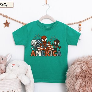 May include: A kelly green short-sleeve t-shirt featuring a graphic print of three spider-themed characters in front of an American flag and fireworks. The word AMERICA is printed in large, colorful, dotted letters at the bottom of the graphic design.