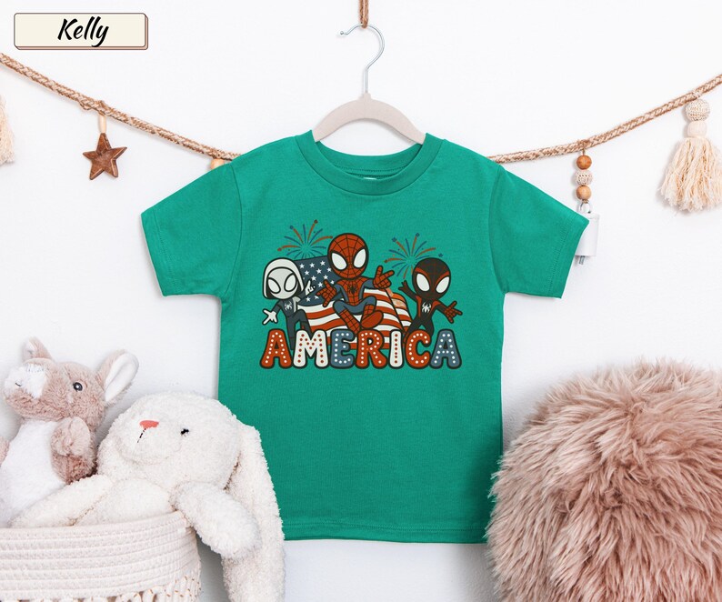 May include: A kelly green short-sleeve t-shirt featuring a graphic print of three spider-themed characters in front of an American flag and fireworks. The word AMERICA is printed in large, colorful, dotted letters at the bottom of the graphic design.