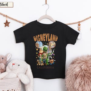 May include: Black t-shirt featuring a Disneyland graphic with the text "Disneyland California" in a vintage style. The shirt showcases various Toy Story characters. The shirt is hanging on a white hanger.