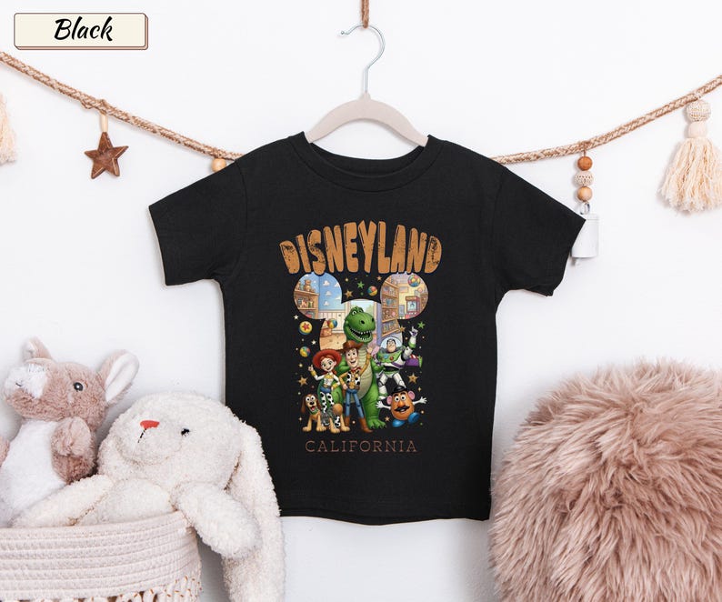 May include: Black t-shirt featuring a Disneyland graphic with the text "Disneyland California" in a vintage style. The shirt showcases various Toy Story characters. The shirt is hanging on a white hanger.